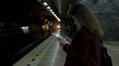 Kız Metro İstasyonu'na ve tren için bekler, akıllı telefon veya tablet kullanarak kırmızı paltolu. Kadın kullanın cep telefonu ve ayakta şehir metro staton.