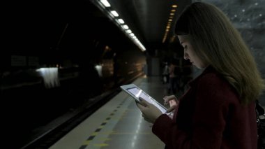 Kız Metro İstasyonu'na ve tren için bekler, akıllı telefon veya tablet kullanarak kırmızı paltolu. Kadın kullanın cep telefonu ve ayakta şehir metro staton.