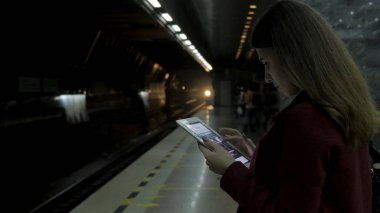 Kız Metro İstasyonu'na ve tren için bekler, akıllı telefon veya tablet kullanarak kırmızı paltolu. Kadın kullanın cep telefonu ve ayakta şehir metro staton.