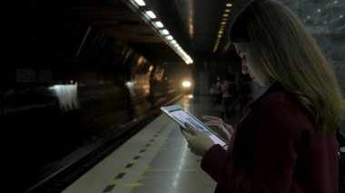 Kız Metro İstasyonu'na ve tren için bekler, akıllı telefon veya tablet kullanarak kırmızı paltolu. Kadın kullanın cep telefonu ve ayakta şehir metro staton.