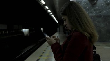 Kız Metro İstasyonu'na ve tren için bekler, akıllı telefon veya tablet kullanarak kırmızı paltolu. Kadın kullanın cep telefonu ve ayakta şehir metro staton.
