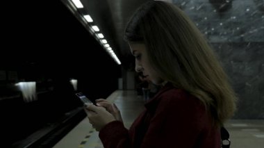 Kız Metro İstasyonu'na ve tren için bekler, akıllı telefon veya tablet kullanarak kırmızı paltolu. Kadın kullanın cep telefonu ve ayakta şehir metro staton.