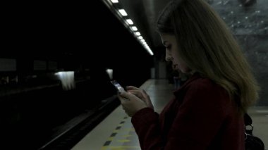 Kız Metro İstasyonu'na ve tren için bekler, akıllı telefon veya tablet kullanarak kırmızı paltolu. Kadın kullanın cep telefonu ve ayakta şehir metro staton.