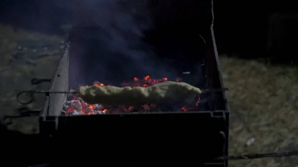 Man placing raw meat onto a hot barbecue to grill using a pair of wooden tongs, close up view over a dark background. Dinner party, barbecue and roast pork at night. Grill background — Stock Photo, Image