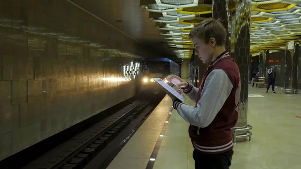 Człowiek za pomocą płytki dotykowej lub smartfona, gdy czeka na pociąg na stacji metra. Samotny młody człowiek z smartphone strzał z profilu na stacji metra z niewyraźne jadącego pociągu w tle. — Zdjęcie stockowe