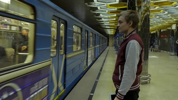 Mies käyttää kosketuslevyä tai älypuhelinta odottaessaan junaa metroasemalla. Yksinäinen nuori mies älypuhelin ammuttiin profiilin metroasemalla sumea liikkuva juna taustalla . — kuvapankkivalokuva