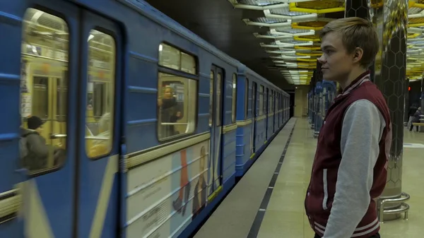 Mies käyttää kosketuslevyä tai älypuhelinta odottaessaan junaa metroasemalla. Yksinäinen nuori mies älypuhelin ammuttiin profiilin metroasemalla sumea liikkuva juna taustalla . — kuvapankkivalokuva