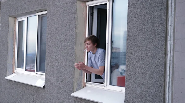 Nuori mies polttaa tupakan keittiön asunnon ikkunasta. Nuori mies polttaa ulos ikkunasta asunnon — kuvapankkivalokuva