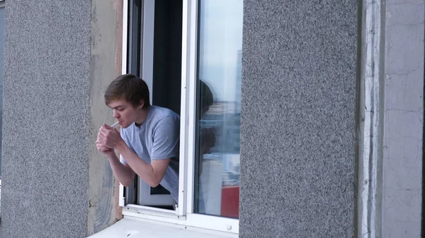 O jovem fuma um cigarro pela janela do apartamento na cozinha. Jovem fuma pela janela do apartamento — Fotografia de Stock