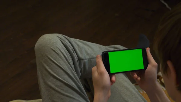 Jovem atraente olhando seus telefones celulares tela verde em branco, enquanto sentado no sofá em casa. Jovem em casa olhando para a tela verde do telefone — Fotografia de Stock