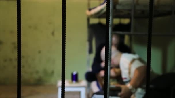 Prisoners wearing prison uniform has lost in thought while lying in bed near bedside table with aluminum dishes in a prison cell. Prisoners behind bars are sitting on the bed — Stock Video