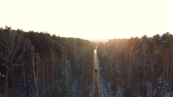 Auto jezdí po silnici v zasněženém lese. Záběry. Paprsky ranního slunce. Letecký pohled. Letecký pohled na zasněžený les s vysoké borovice a silnici s autem v zimě. Pohled shora na zimní silnici — Stock fotografie
