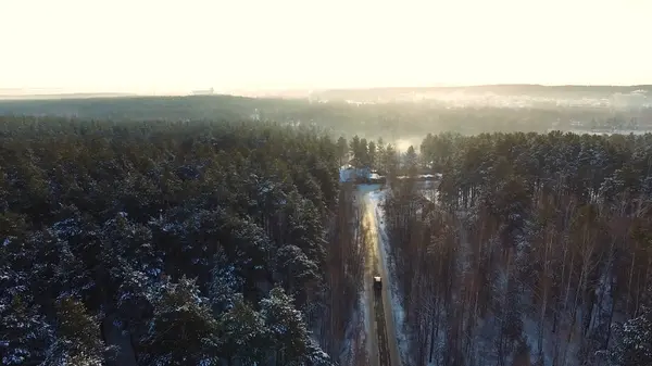 Auto rijdt over de weg in het besneeuwde bos. Beeldmateriaal. Stralen van de ochtendzon. Luchtfoto. Luchtfoto van een besneeuwde forest met hoge dennen en weg met een auto in de winter. Bovenaanzicht van winter weg — Stockfoto