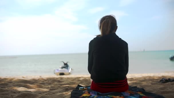 Widok dziewczyny w Bluza z kapturem, siedząc na plaży z tyłu i oglądać w tle morze, Błękitne niebo i morze. Klip. Młoda kobieta w sportowej siedzi na plaży — Wideo stockowe