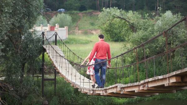 Unga älskare på en repbro över floden. Ett par vandrare korsar en sväng brygga. Klipp. Ungt par i kärlek gå på hängande träbro över en sjö i skogen. — Stockfoto