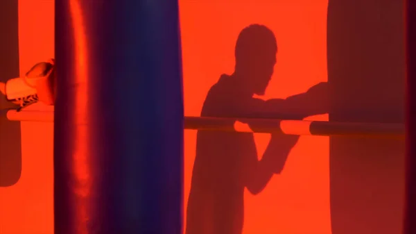 Silhouette or shadow of a boxer punching a punching bag close up, red light background. Silhouette of young male boxer hitting punching bag on red background. Man athlete standing and boxing.