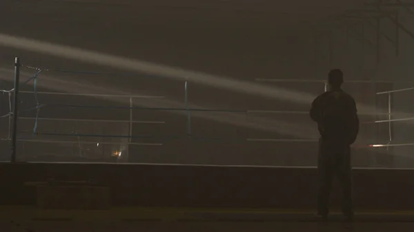 Fighter man silhouette dark. Mma fighter silhouette on dark ring. Boxer stands on stage before fight. Boxer silhouette in smoke at dark studio. Rear view on man near to the Boxing ring