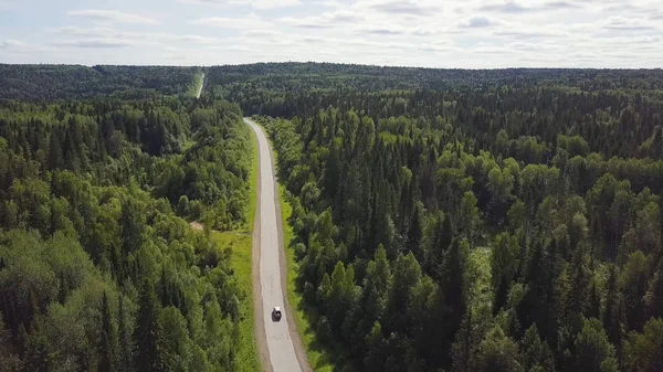 Aerial view of white car driving on country road in forest. Aerial view flying over old patched two lane forest road with car moving green trees of dense woods growing both sides. Car driving along