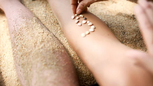 As mãos femininas reúnem o modelo de conchas e areia, perto. Vídeo. Pedaços de âmbar e conchas em mãos femininas em um fundo de areia. Menina brinca com conchas e areia sentada na praia . — Fotografia de Stock