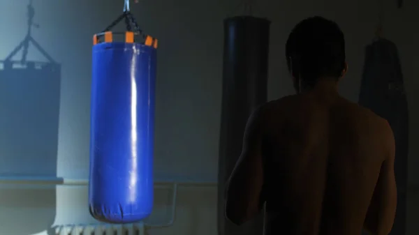 Back view of muscular male boxer punching bag in boxing gym. Fighter blows jab cross hook series, working his hands with boxing gloves. Sporty man coming closer to the boxing bag and starting to kick — Stock Photo, Image