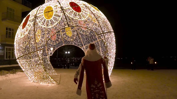 Rear view of Santa Clous coming to the Christmas lights decorations in the street at night, winter holidays concept. Clip. Amazing spherical construstion covered with garlands. — Stock Photo, Image