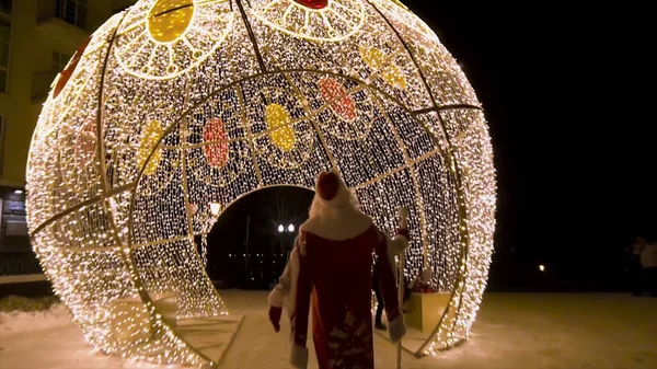Rear view of Santa Clous coming to the Christmas lights decorations in the street at night, winter holidays concept. Clip. Amazing spherical construstion covered with garlands. — Stock Photo, Image