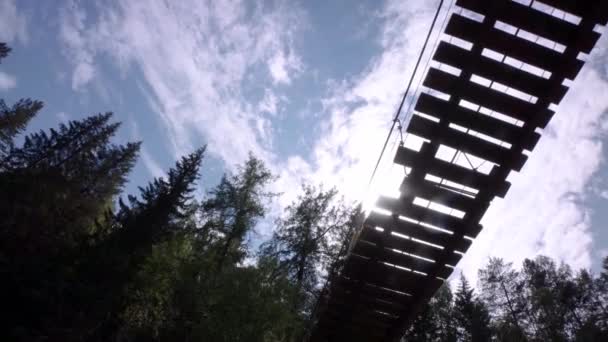 Botten syn på människan på hängbro. Lagerbilder. Man går på trä hängbro på bakgrund av gran toppar och blå himmel med sol — Stockvideo