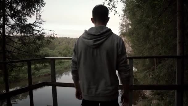 Hombre joven se encuentra en el fondo del río, bosque verde y cielo nublado. Imágenes de archivo. Hermosa plataforma de madera para turistas que admiran la vista del paisaje forestal con el río que fluye — Vídeo de stock