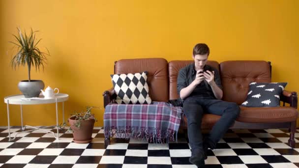 O tipo joga ao telefone sentado no sofá. Imagens de stock. Jovem joga entusiasticamente telefone sentado no interior elegante. Jovem passa o tempo de lazer jogos no telefone — Vídeo de Stock