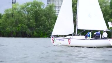 Rüzgarlı havada, yeşil ağaçlar ve bulutlu arka planda beyaz teknelerle nehrin aşağısına yelken açan insanlar. Spor anlayışı. - Sanat. Küçük yatlar nehir dalgaları üzerinde yüzüyor..