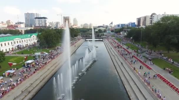 Aerial view of the Yekaterinburg city embankment and the Iset river with the crowd of people walking nearby. Action. Top view of the river with fountains. — ストック動画