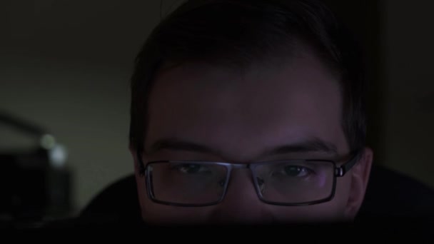 Detailní záběr mladého obchodníka, jak se dívá na monitor, když sedí v temné místnosti. Záběry ze skladu. Muž s brýlemi při pohledu na obrazovku počítače. — Stock video