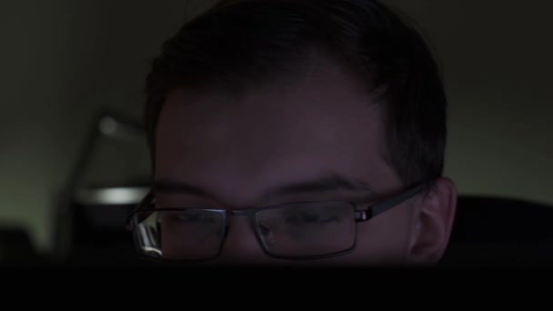 Κοντινό πλάνο του νεαρού επιχειρηματία που κοιτάζει την οθόνη ενώ κάθεται στο σκοτεινό δωμάτιο. Στικ. Άντρας με γυαλιά που κοιτάζει την οθόνη του υπολογιστή. — Αρχείο Βίντεο