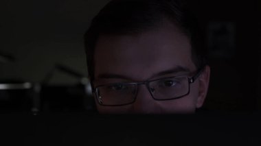Karanlık odada dizüstü bilgisayar kullanan odaklanmış ve düşünceli bir adam kodlayıcı, programcı ya da geliştirici. Stok görüntüleri. Yüzünün yarısı perdenin arkasında gözlüklü genç adama yaklaş..