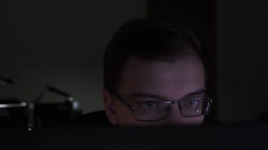 Karanlık odada dizüstü bilgisayar kullanan odaklanmış ve düşünceli bir adam kodlayıcı, programcı ya da geliştirici. Stok görüntüleri. Yüzünün yarısı perdenin arkasında gözlüklü genç adama yaklaş..