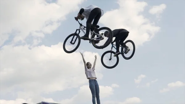 Rosja - Moskwa, 08.15.2019: dwóch zawodników skaczących z trampoliny na tle błękitnego zachmurzonego nieba. Akcja. Dwóch jeźdźców Bmx robi sztuczki i przybija piątkę dziewczynie. — Zdjęcie stockowe