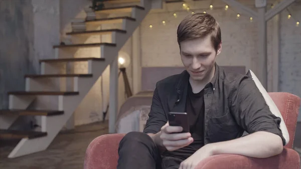 Joven hombre usando un teléfono inteligente sentado en el sillón en casa en el apartamento dúplex con luces de guirnalda detrás de él. Imágenes de archivo. Hombre con teléfono celular relajante en la habitación espaciosa . — Foto de Stock