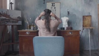 Masasında oturup saçlarına dokunan esmer bir kadının arka görüntüsü. Stok görüntüleri. Gri duvarlı arka planda sanat atölyesinde çalışan kadın..