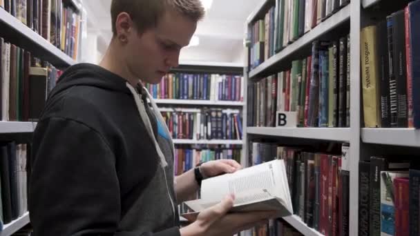 Duitsland - Berlijn, 09.10.19: Jongeman in een boekhandel met een geopend boek in zijn handen met andere boekenplanken op de achtergrond, onderwijs en leesconcept. Voorraadbeelden. Man in de bibliotheek of — Stockvideo