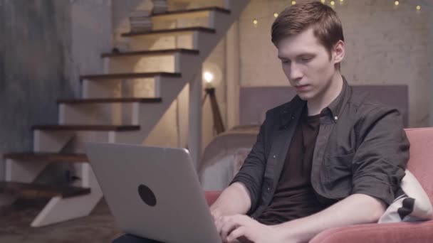 Jongeman werkt met laptop in fauteuil, op duplex flat interieur achtergrond, freelance concept. Voorraadbeelden. Man typen op zijn computer. — Stockvideo