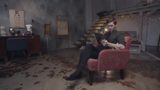 Portrait de jeune homme portant une chemise sombre regardant sa tablette numérique dans une pièce stylisée ou un atelier d'art. Images d'archives. Homme utilisant Ipad assis dans le fauteuil rouge . — Video