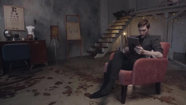 Beau jeune homme dans un fauteuil rouge à l'aide d'une tablette numérique. Images d'archives. Homme regardant son appareil dans un authentique studio d'art duplex aux murs gris . — Video