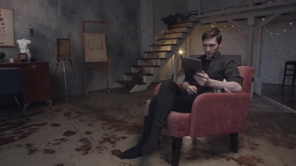 Knappe jongeman in een rode fauteuil met een digitale tablet. Voorraadbeelden. Man op zoek naar zijn apparaat in authentieke duplex art studio met grijze muren. — Stockvideo