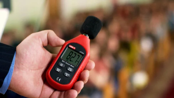 Close up of a man hand holding a red sound level meter and analyzer on clapping audience background. Stock footage. Man measuring the noise level on blurred crowd background. — Stock Photo, Image