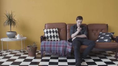 Genç adam, siyah beyaz zemin ve sarı duvarlı bir odada deri koltukta otururken cep telefonu kullanarak mesaj yolluyor. Stock Video. Modern teknoloji kavramı.