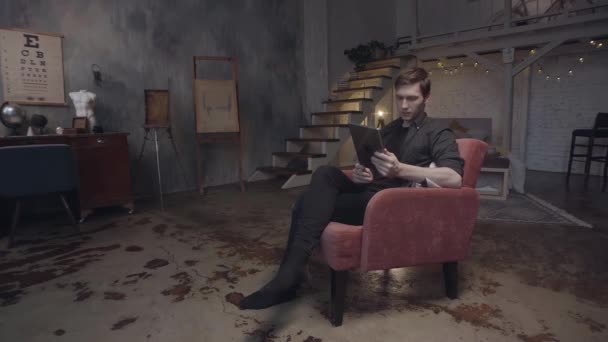 Portrait de jeune homme portant une chemise sombre regardant sa tablette numérique dans une pièce stylisée ou un atelier d'art. Images d'archives. Homme utilisant Ipad assis dans le fauteuil rouge . — Video