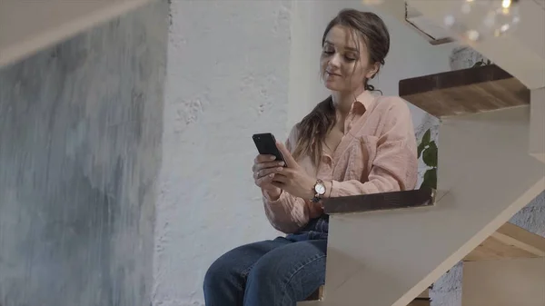 Stilvolle Frau, die auf Sternen in ihrem Häuschen sitzt, während sie Nachrichten liest und lächelt, Online-Kommunikationskonzept. Archivmaterial. hübsche Frau mit ihrem Handy zu Hause. — Stockfoto