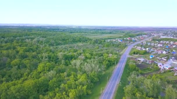 青い曇りの空の背景にある小さな町への緑の森を介して、道路の空中ビュー。アート。新鮮な夏の木や道路のトップビュー. — ストック動画