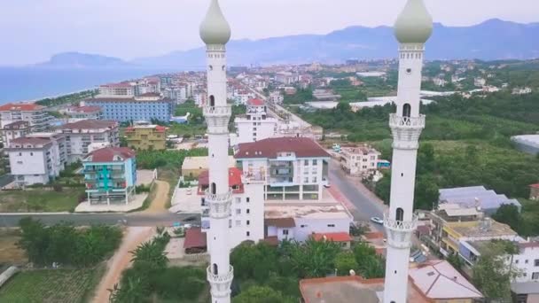 Widok z lotu ptaka dwóch minaretów meczetu otoczonego ulicami miasta z domami, parkami i zielonymi drzewami. Sztuka. Latanie przez minarety nad letnim miastem. — Wideo stockowe