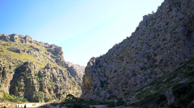 Yeşil çayırlarla kaplı dik yamaçlı iki dağ arasındaki patika. - Sanat. Mavi arka planda, nefes kesici kayaların yanında yürüyen insanlar..